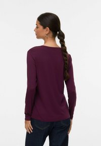 Lange mouwen, ronde hals top in diep bordeauxkleurige stof, gladde textuur. Gecombineerd met donkere denim. Zichtbare vlechtstijl en grote oorbellen.
