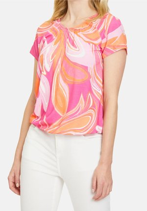 Donna che indossa una camicetta a maniche corte con motivo a vortice rosa, arancione e bianco, abbinata a pantaloni bianchi, mostrata dal collo ai fianchi.