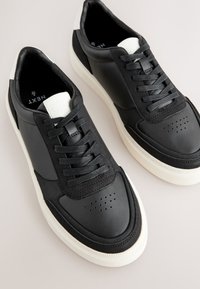 Baskets en cuir noir avec un design texturé, fermeture à lacets, bout perforé, et semelle en caoutchouc blanche contrastante.