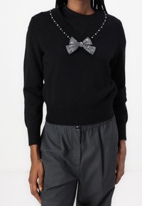 Czarny sweter z dekoltem w serek, ozdobiony rhinestones wokół kołnierza oraz błyszczącą kokardą z przodu na środku. Noszony na szarych spodniach.