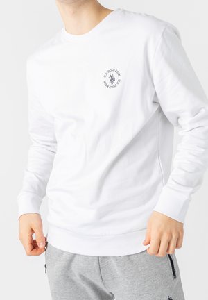 Mężczyzna w białej koszulce z długim rękawem U.S. Polo Assn. z logo na piersi, w połączeniu z jasnoszarymi spodniami dresowymi z kieszeniami na suwak.
