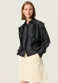 Black ruffled blouse with a collared neck and button closure, featuring puffed sleeves and front pockets. Worn over a light beige skirt.