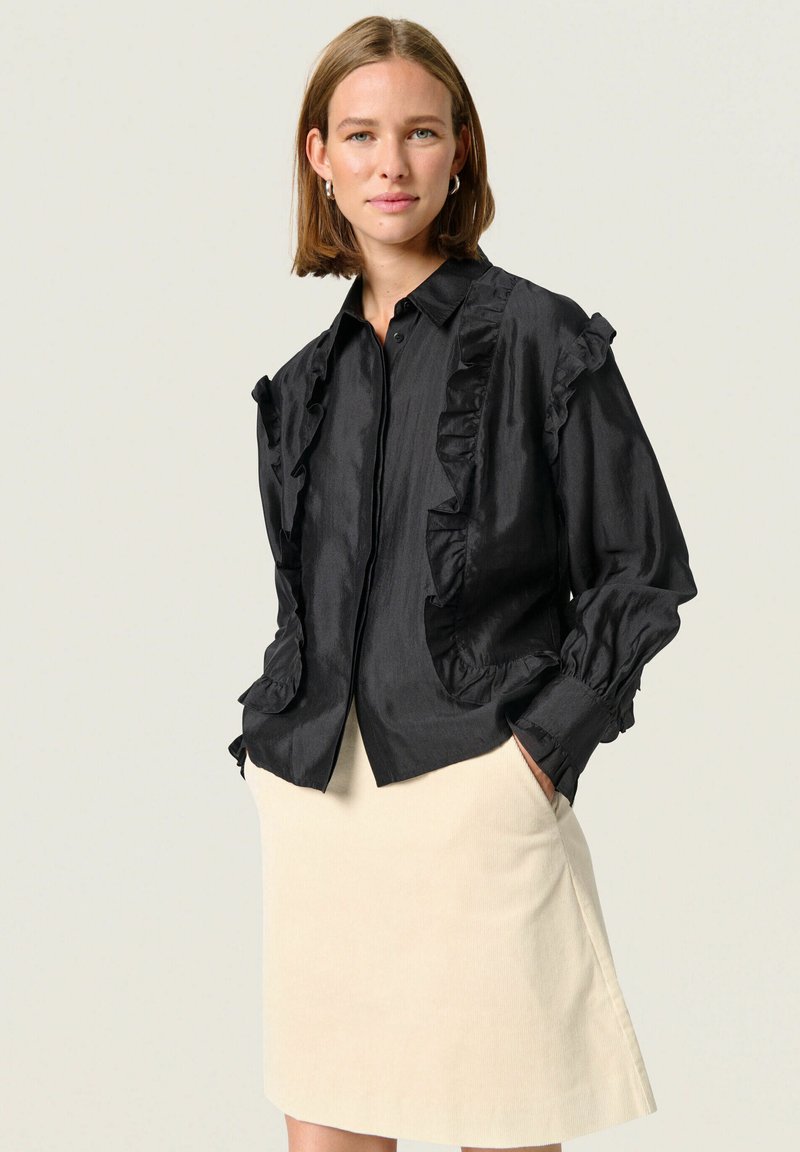 Zwarte gerimpelde blouse met een kraag en knoopsluiting, voorzien van pofmouwen en voorzakken. Draag over een lichtbeige rok.