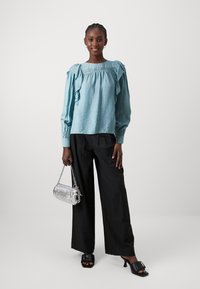 Blusa azzurra chiara in tessuto texture con maniche a volants, abbinata a pantaloni neri a gamba larga e una borsa metallica argento. Mules nere con punta aperta.