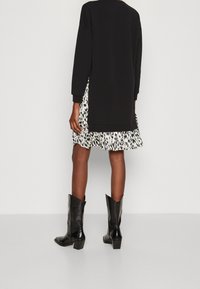 Schwarzes Langarmoberteil mit lässigem Schnitt, kombiniert mit einem gestuften weißen Kleid mit abstraktem schwarzen Muster; schwarze Stiefeletten mit Absatz runden den Look ab.