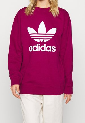 Person, die einen magentafarbenen Adidas Sweatshirt mit großem weißem Trefoil-Logo und weißen Hosen trägt, vor einem einfarbigen weißen Hintergrund stehend.