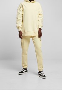 Osoba ubrana w kremowy, oversize'owy hoodie, pasujące joggery cargo oraz czarno-białe sneakersy, stojąca na tle jednolitego, jasnoszarego tła.