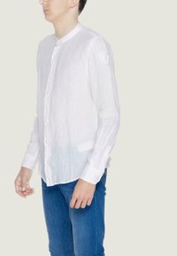 Camicia bianca in lino con colletto rialzato, chiusura con bottoni e una sfumatura graduale che passa a un blu chiaro sull'orlo. Presenta maniche lunghe e un'etichetta con logo.