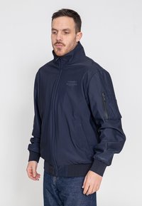 Marine Bomberjacke mit hohem Kragen, Frontreißverschluss, Seitentaschen und einer aufgesetzten Tasche am linken Arm. Hergestellt aus leichtem, glattem Stoff.