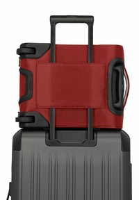 Valigia rigida nera con texture a righe, sopra la quale è posizionato un sacco di tessuto rosso fissato con una cinghia e dotato di accessori e maniglie nere.