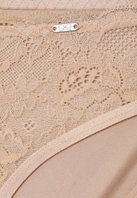 Beige kanten slipjes met een bloemenpatroon, voorzien van een gladde stoffen basis en een klein metalen label met een gegraveerd ontwerp.