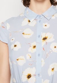 Ljusblommig klänning i blått med korta ärmar, prydd med vita blommor och gyllene detaljer, knäppning fram och krage.