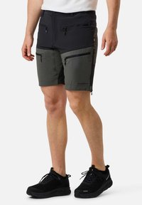 Uomo che indossa pantaloncini con cerniera bicolore nero e verde oliva e scarpe da ginnastica nere, in piedi davanti a uno sfondo bianco.