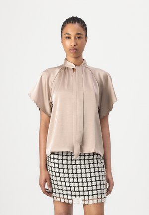 Femme portant un chemisier en satin beige avec des manches volantées et des bords festonnés, associé à une mini-jupe en dentelle florale noire et blanche.