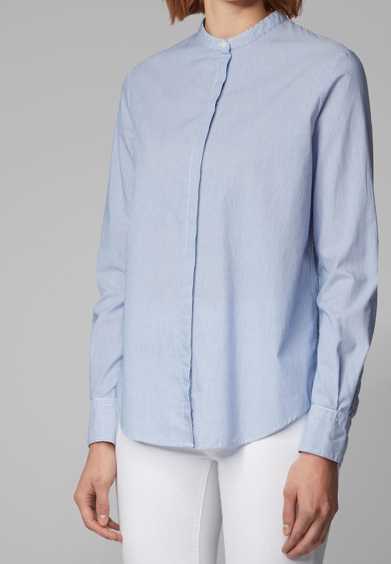 Femme portant une chemise bleu clair à fines rayures avec col mandarin et pantalon blanc sur fond gris uni.