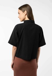 Korte zwarte cropped shirt met korte mouwen en een gestructureerde stof, voorzien van een klassieke kraag en een rechte zoom. Draag het over een bruine rok.