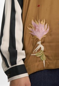Pruun kangast blouse, millel on tikitud muster, kus on kujutatud figuur ja roosa lill. Triibulised mustad ja kreemjad varrukad, sile tekstuur.