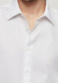 Chemise blanche boutonnée avec un tissu texturé, dotée d'un col et d'une rangée de boutons transparents le long de la patte de boutonnage. Vue en gros plan.