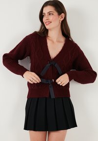 Burgundy knitted cardigan featuring a V-neck, textured cable pattern, and navy ribbon tie closure at the front. Paired with a pleated black skirt.