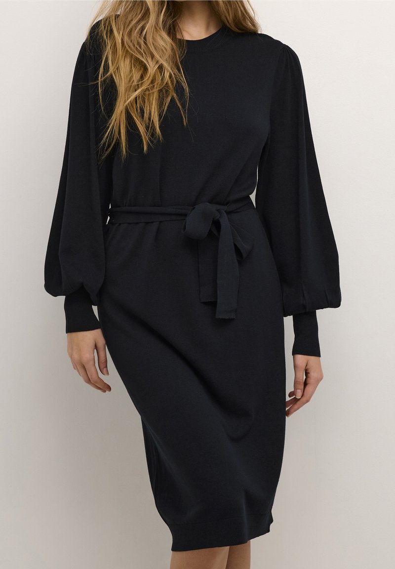 Femme portant une robe midi noire à manches longues bouffantes avec une ceinture nouée à la taille, debout devant un fond clair uni.