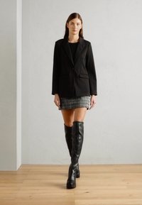 Black tailored blazer over a black top, paired with a houndstooth mini skirt and knee-high black leather boots, set against a neutral background.