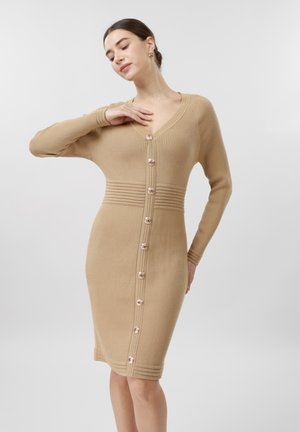 Femme portant une robe en maille côtelée beige ajustée à manches longues avec une patte de boutonnage à l'avant, posant avec une main près de son cou.