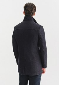 Czarny płaszcz w wzory z dopasowanym krojem. Posiada wysoki kołnierz, wyraźne ramiona oraz centralne rozcięcie z tyłu dla swobody ruchów.