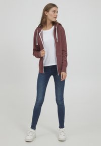 Haut zippé bordeaux avec cordon blanc, porté sur un t-shirt blanc. Associé à un jean skinny bleu et des baskets blanches.