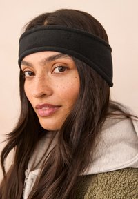 Crna traka za glavu od flisa s dva vidljiva šava, nošena preko duge tamne kose. Mekana tekstura na svijetloj pozadini.