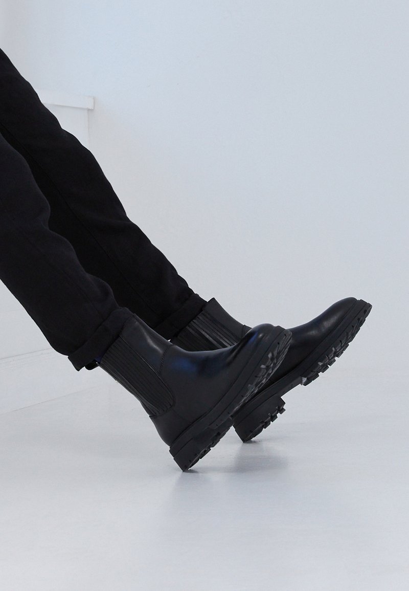 Persona che indossa stivali Chelsea in pelle nera con lato elastico e pantaloni neri, gambe sollevate contro uno sfondo chiaro e semplice.