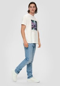 Biała bawełniana koszulka z kwadratowym wzorem graficznym w kolorach fioletowym i zielonym. Noszona z jasnoniebieskimi dżinsami i białymi sneakersami.