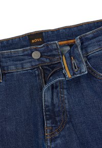 Tmavě modré džíny z denimu s kontrastním oranžovým prošitím, kovovým knoflíkem a zipovým zapínáním. Pas obsahuje černou nálepku s nápisem "BOSS."