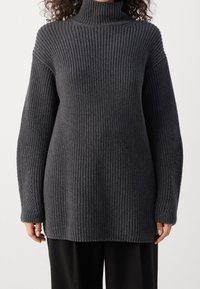 Tlustý tmavě šedý svetr s vysokým límcem, žebrovanou strukturou, spadlými rameny a dlouhými rukávy, sahajícími do poloviny stehen.