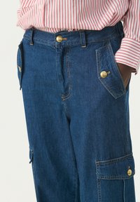 Pantalones cargo de mezclilla en azul oscuro, con herrajes dorados, múltiples bolsillos y un diseño de pierna recta. Tejido texturizado con detalles de costura.
