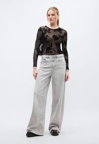 Μαύρο μακρυμάνικο τοπ με σχέδιο από λουλουδάτη δαντέλα, συνδυασμένο με φαρδιά ανοιχτό γκρι τζιν παντελόνια και μαύρα παπούτσια πλατφόρμας. Συμπεριλαμβάνεται μικρή μαύρη τσάντα χειρός.