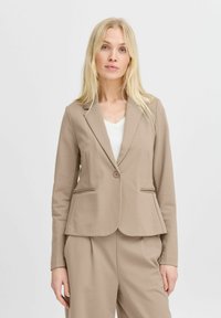 Béžový látkový blazer s přiléhavým střihem, jednobotonovým zapínáním a dvěma bočními kapsami. Nosený přes bílý top, má klasický klopový límec.