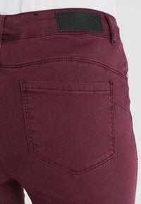 Los jeans de mezclilla burdeos presentan una vista trasera con un parche de cuero negro, detalles de costura visibles y un diseño estándar de bolsillo trasero.