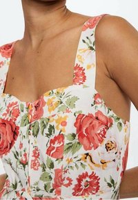 Bloemen crop top met een vierkante halslijn, brede bandjes en knoopdetails. Gemaakt van een lichte stof met een levendig rood en geel bloemenpatroon.