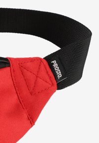 Czerwony produkt z tkaniny z czarnym paskiem, z trójkątnym przeszyciem. Etykieta "PROSTO" widoczna na pasku w miejscu łączenia.