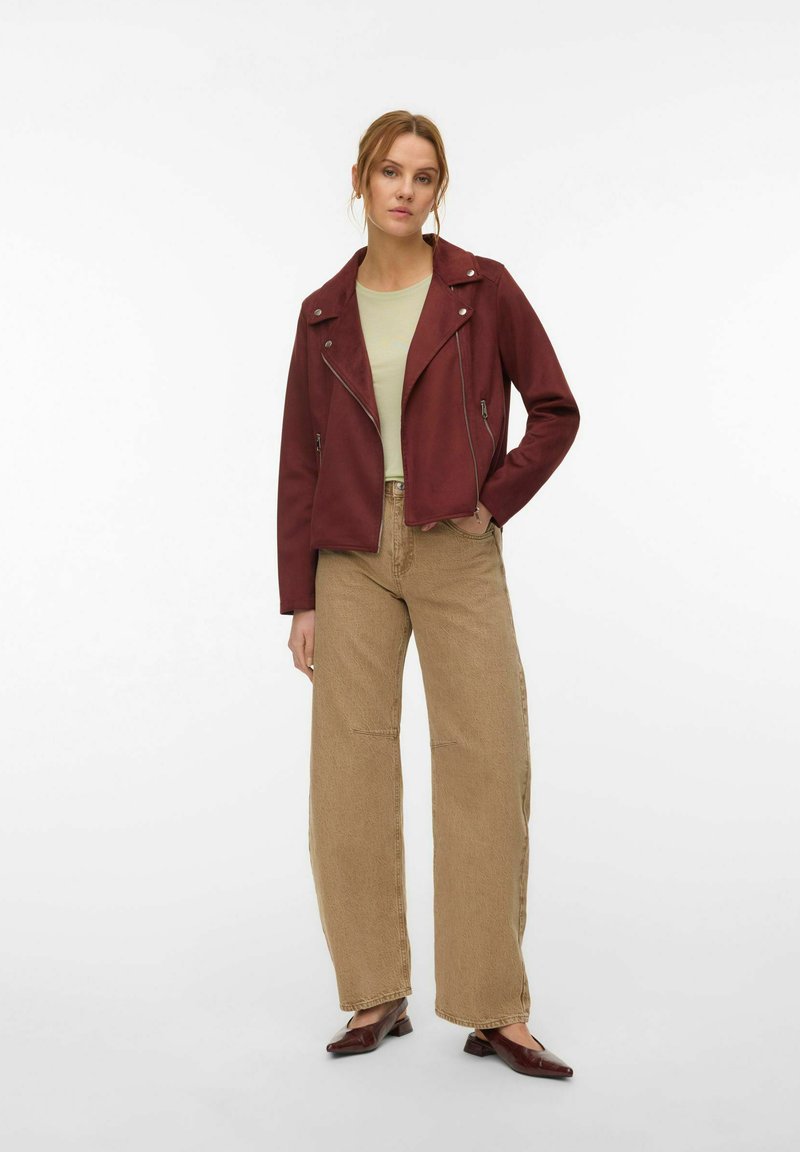 Femme portant une veste zippée bordeaux, un haut vert clair, un pantalon beige à jambes larges et des chaussures bordeaux à petit talon, debout avec une main dans la poche.