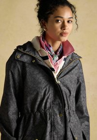 Čierny kabát s kvetinovým vzorom, so sťahovacím pásom, zipsovým zapínaním a zlatými doplnkami. Nosí sa cez ružovú košeľu s golierom a vzorovaný šál.