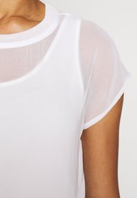 Bílý, průsvitný top s krátkými rukávy a kulatým výstřihem. Látka působí lehce a má hladkou, mírně průhlednou strukturu.