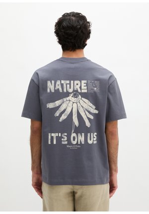 Mężczyzna w szarej koszulce z wyblakłym nadrukiem kwiatowym i napisem „NATURE IT'S ON US” na plecach, stojący na jednolitym białym tle.