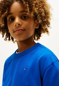 T-shirt di cotone blu con collo a giro, con un piccolo logo rosso, bianco e blu sul lato sinistro del petto. Capelli ricci parzialmente visibili.