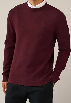 Homme portant un pull ras du cou texturé rouge foncé sur une chemise blanche à col, avec une main dans la poche d'un pantalon noir.