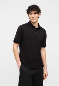 Νεαρός άνδρας με σγουρά μαλλιά που φοράει ένα μαύρο πόλο με κοντά μανίκια και πατιλέτα με κουμπιά, όρθιος με το ένα χέρι στην τσέπη μπροστά σε λευκό φόντο.