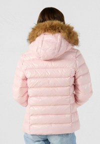 Persona que lleva una chaqueta puffer rosa claro con capucha de piel sintética, vista desde atrás, combinada con jeans azul claro.