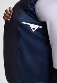 Innenansicht eines schwarzen Anzugsakkos mit glattem marineblauem Futter, weißem Taschenbesatz und Knopf. Der Stoff wirkt strukturiert, strapazierfähig und gut verarbeitet.