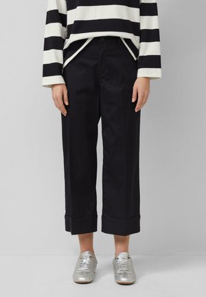 Osoba ubrana w czarne szerokie, krótkie spodnie, srebrne metaliczne tenisówki oraz białą koszulę w czarne paski z długim rękawem, z rękami swobodnie opuszczonymi wzdłuż ciała.