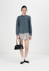 Kvinne iført en blå knappet bluse og grå plisserte skjørt, som holder en svart håndveske, med hvite sokker og svarte ballerinasko.